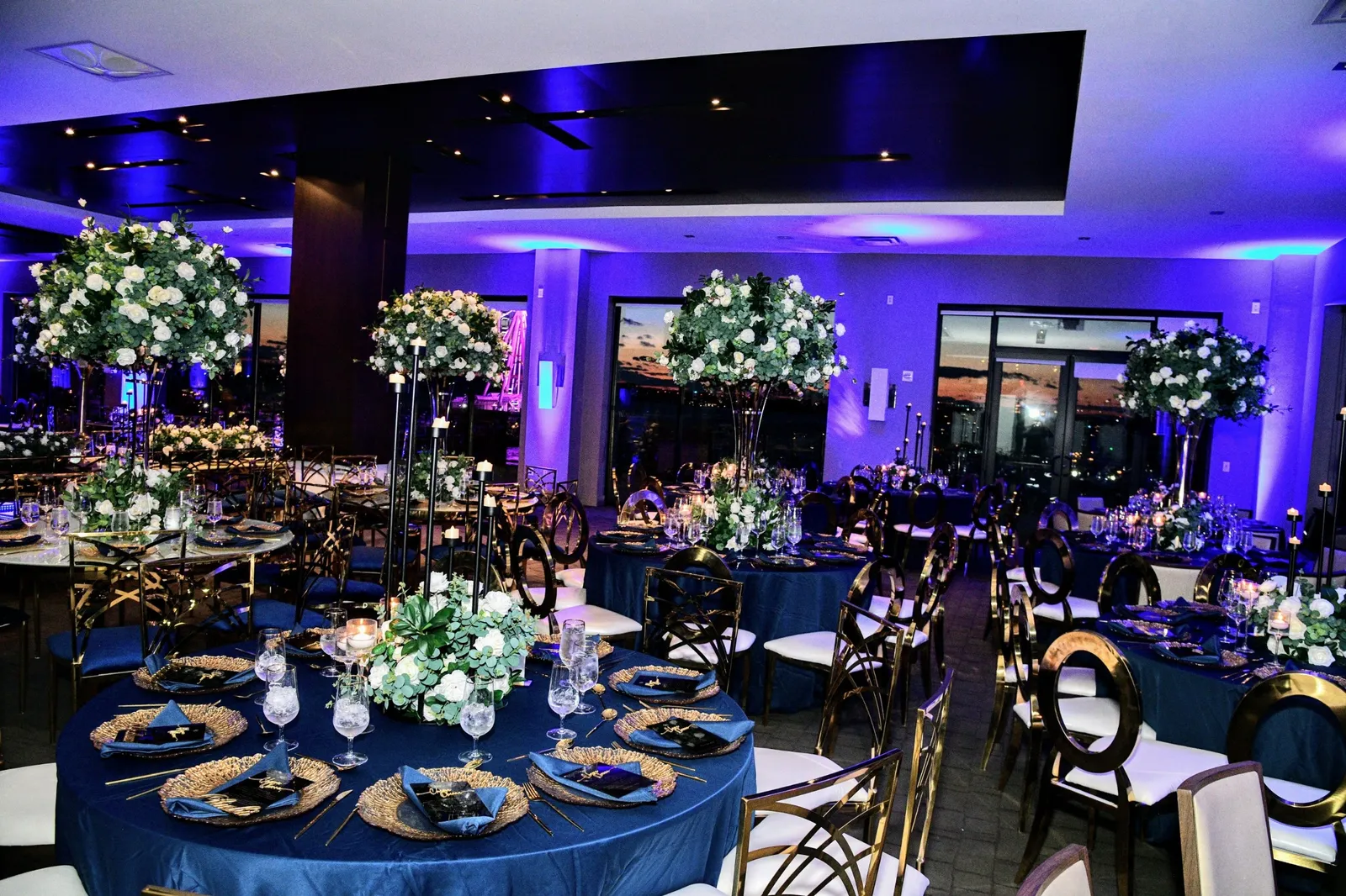 Reception room at sunset with tall white rose centerpieces, navy tables and the waterfront through the windows